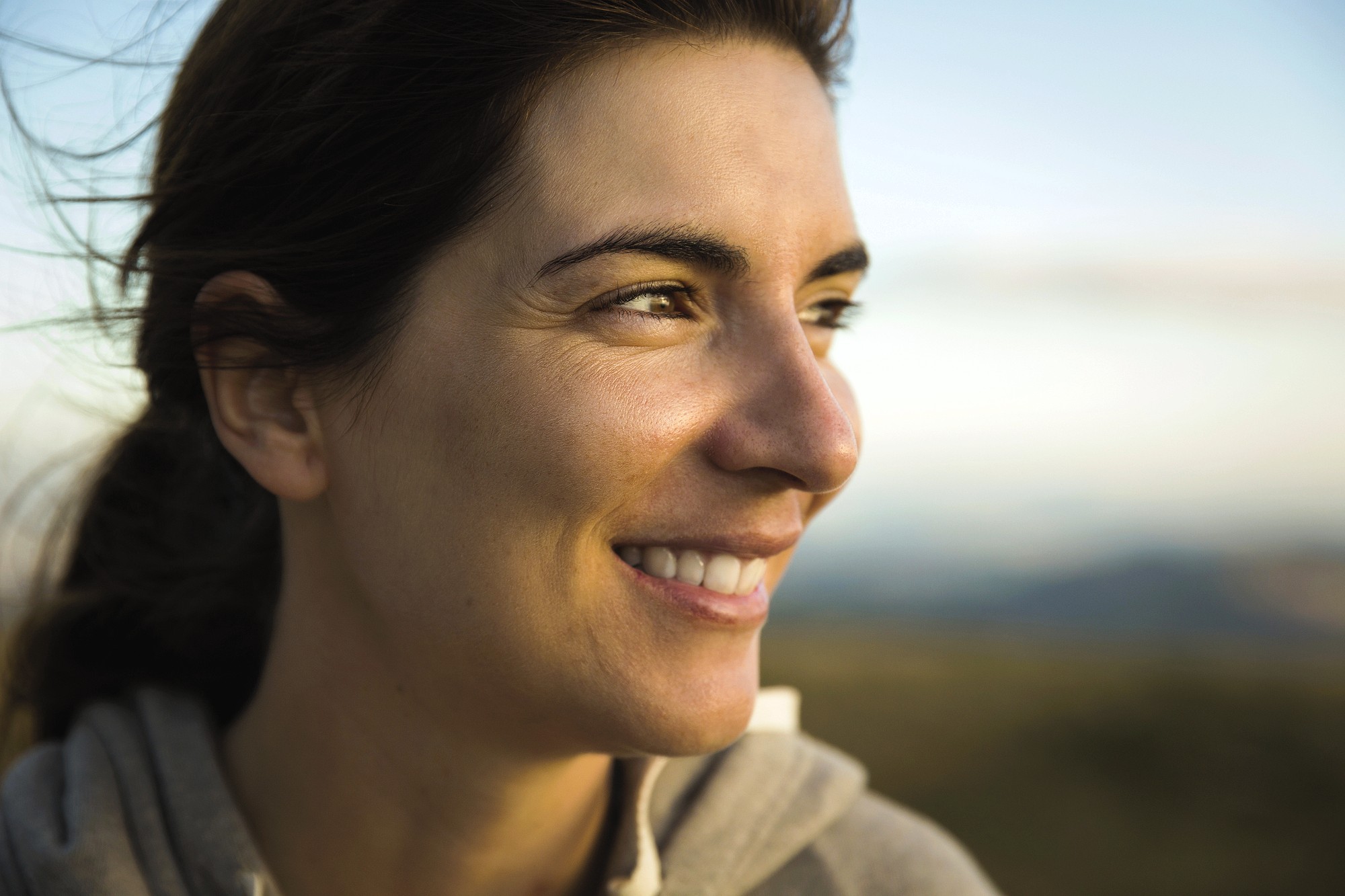 A person with long dark hair smiles while gazing to the side. The background is a blurred natural setting, suggesting an outdoor location. The lighting is warm and soft, casting a gentle glow on the person's face.