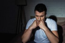 A man sits on a bed in a dimly lit room, wearing a grey t-shirt. He appears deep in thought, with his hands clasped under his chin, looking downward. The mood is contemplative and somber.