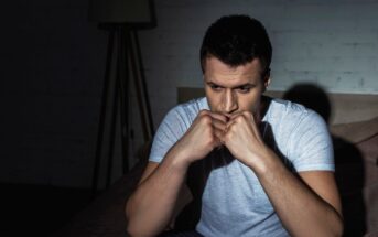A man sits on a bed in a dimly lit room, wearing a grey t-shirt. He appears deep in thought, with his hands clasped under his chin, looking downward. The mood is contemplative and somber.