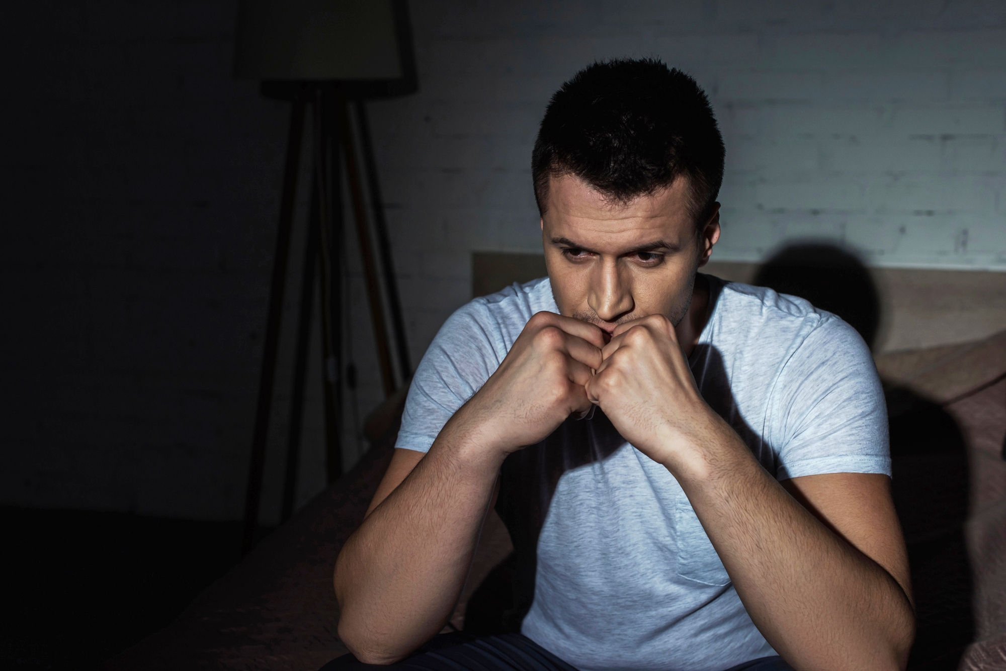 A man sits on a bed in a dimly lit room, wearing a grey t-shirt. He appears deep in thought, with his hands clasped under his chin, looking downward. The mood is contemplative and somber.