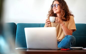 A woman with curly hair and glasses sits cross-legged on a couch, smiling while holding a mug. An open laptop is in front of her on a coffee table, with a notebook and pen nearby.