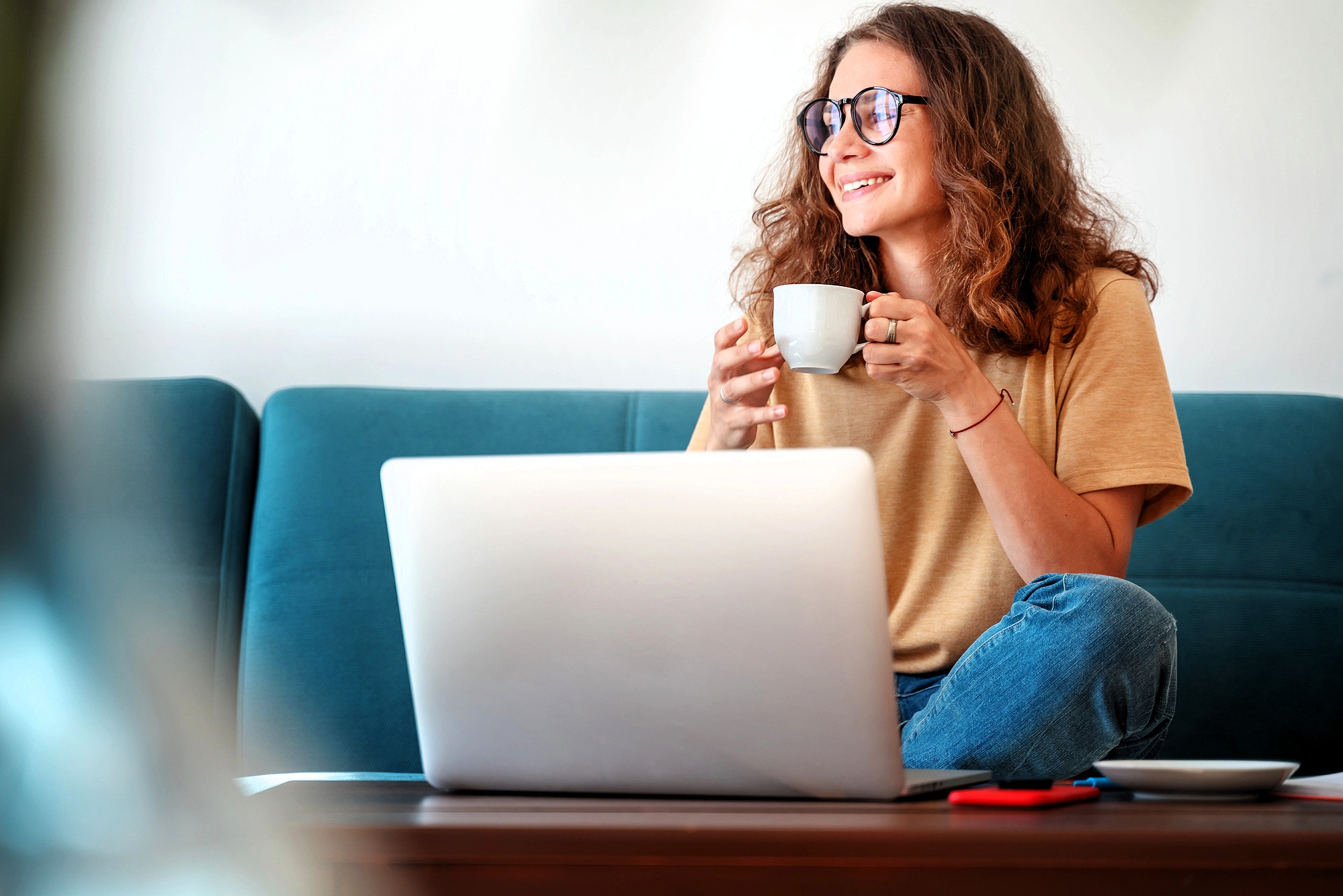 A woman with curly hair and glasses sits cross-legged on a couch, smiling while holding a mug. An open laptop is in front of her on a coffee table, with a notebook and pen nearby.
