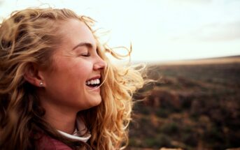 A young woman with long, wavy blonde hair smiles joyfully with her eyes closed outdoors. The wind blows her hair, and a blurred landscape stretches out behind her under a bright sky.