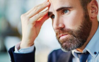 A close-up of a man in a suit with a beard, looking thoughtful and concerned, resting his forehead on his hand.