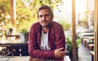 A man with short brown hair and a beard, wearing a maroon shirt over a white t-shirt, sits at a wooden outdoor table, smiling slightly. The background has plants and warm, natural light.