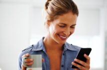 A woman with light brown hair in a bun smiles while looking at her smartphone. She is wearing a blue shirt and holding a gray and white mug in her other hand. The background is softly blurred.