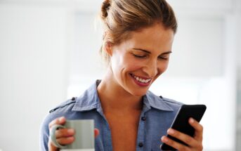 A woman with light brown hair in a bun smiles while looking at her smartphone. She is wearing a blue shirt and holding a gray and white mug in her other hand. The background is softly blurred.