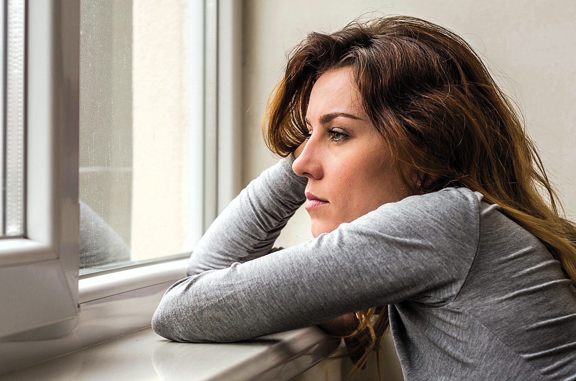 A woman with long brown hair and a gray shirt leans on a windowsill, gazing thoughtfully outside. She's resting her chin on her arm, appearing contemplative and serene, with natural light illuminating her face.
