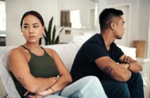 A woman and a man sit on a couch with arms crossed, facing away from each other, appearing upset. The woman is in a green top, and the man in a blue shirt. The room is bright and modern.