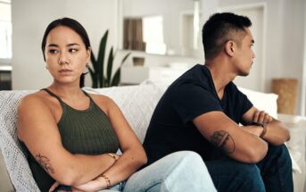 A woman and a man sit on a couch with arms crossed, facing away from each other, appearing upset. The woman is in a green top, and the man in a blue shirt. The room is bright and modern.