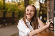 A young woman with long light brown hair sits at an outdoor table, smiling and looking to the side. She is wearing a white shirt and holding a glass jar. Trees and a street are visible in the blurred background.