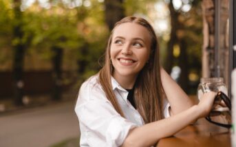 A young woman with long light brown hair sits at an outdoor table, smiling and looking to the side. She is wearing a white shirt and holding a glass jar. Trees and a street are visible in the blurred background.