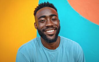 A smiling man with a beard wearing a light blue shirt stands in front of a colorful wall with yellow, blue, and orange sections.