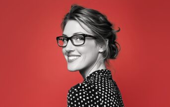 A smiling woman with glasses and a black-and-white polka dot blouse looks over her shoulder against a solid red background.