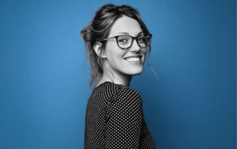 Woman with glasses and long hair tied back, smiling and looking over her shoulder. She is wearing a black and white polka dot top, with a blue background. The image is mostly black and white except for the background.