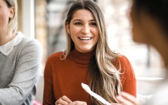 A woman with long brown hair wearing an orange sweater smiles while sitting at a table and talking to two other people in a bright indoor setting.
