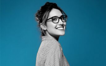 Woman in glasses smiling, looking over her shoulder against a solid blue background. She has dark hair tied back and is wearing a light-colored shirt with small polka dots. The image is mostly black and white except the background.