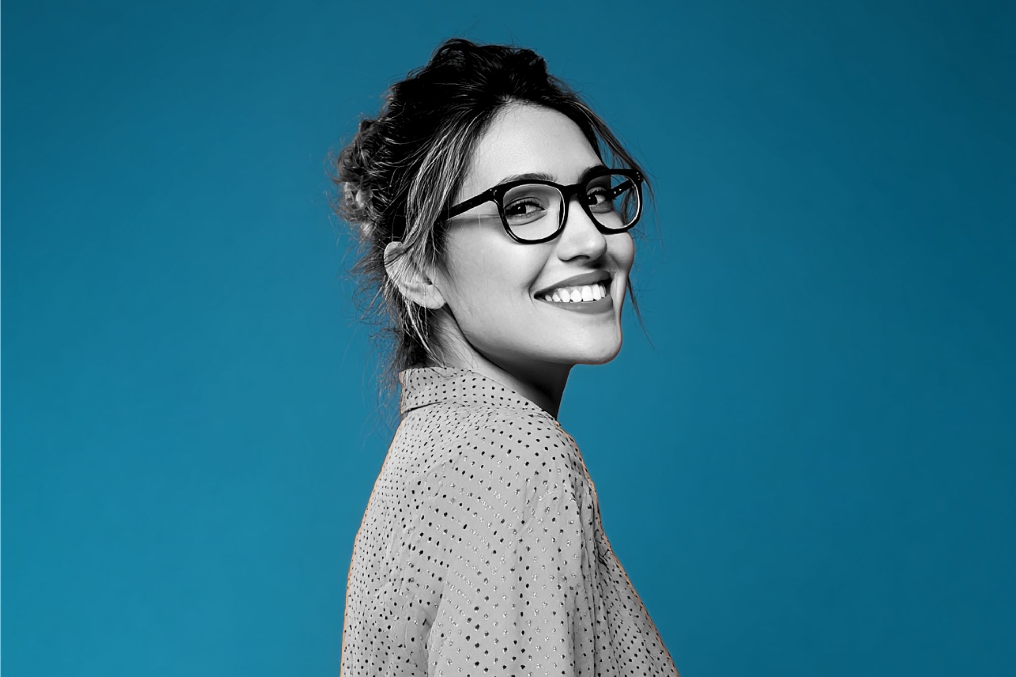 Woman in glasses smiling, looking over her shoulder against a solid blue background. She has dark hair tied back and is wearing a light-colored shirt with small polka dots. The image is mostly black and white except the background.