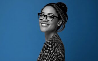 A smiling woman with glasses and her hair in a bun, viewed from the side against a blue background.