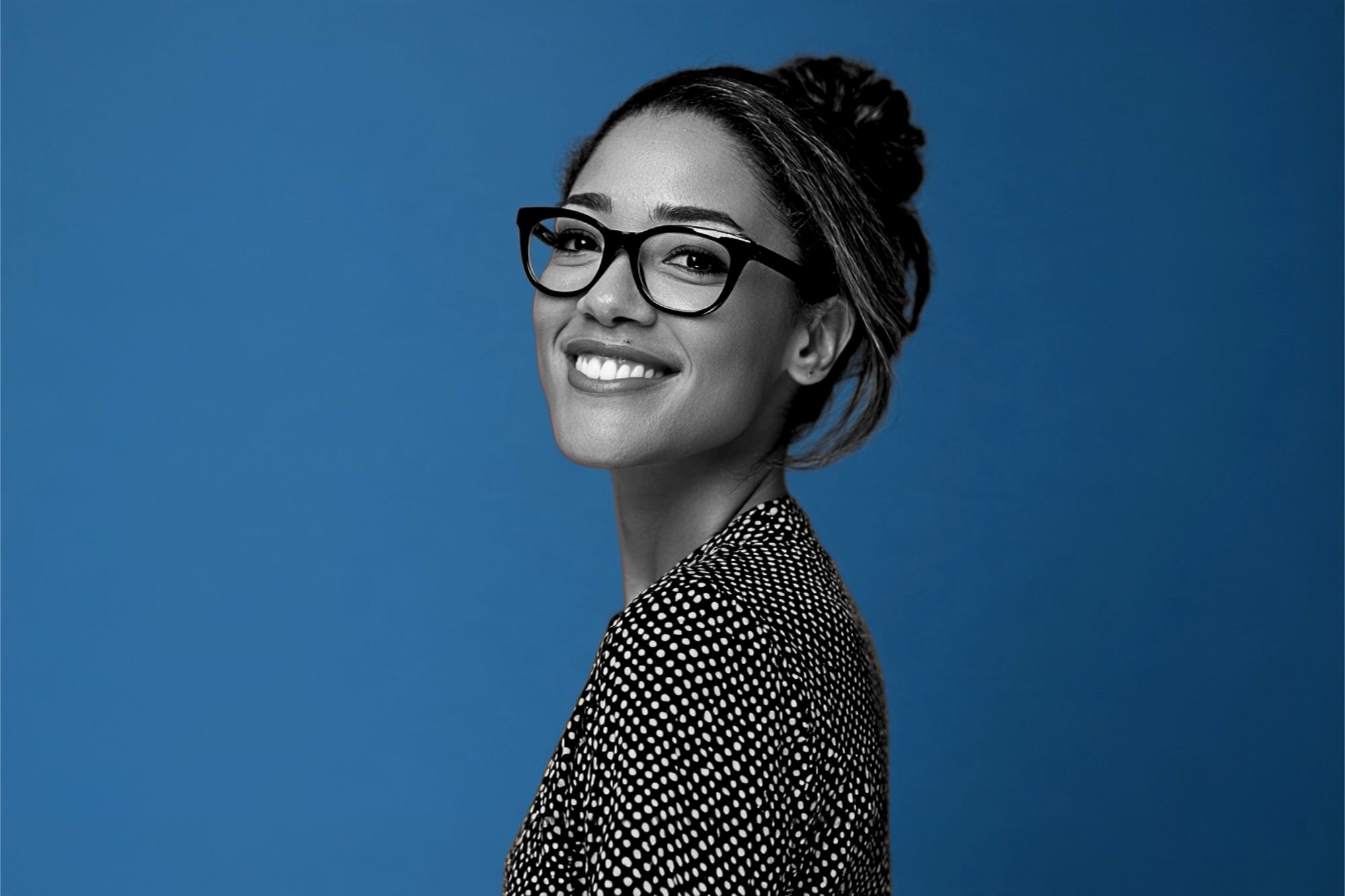 A smiling woman with glasses and her hair in a bun, viewed from the side against a blue background.