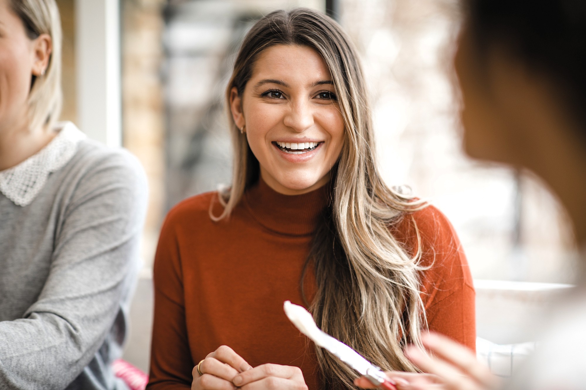 A woman with long brown hair wearing an orange sweater smiles while sitting at a table and talking to two other people in a bright indoor setting.