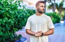 A man with a beard wearing a light-colored t-shirt stands outdoors near leafy greenery, looking to his right with his hands clasped in front of him. Sunlight filters through the trees in the background.
