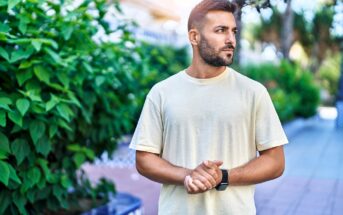 A man with a beard wearing a light-colored t-shirt stands outdoors near leafy greenery, looking to his right with his hands clasped in front of him. Sunlight filters through the trees in the background.