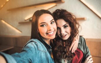 Two people with long hair smile at the camera while taking a selfie indoors. One wears a denim jacket, and the other has a green jacket. The background features angled wooden shelves on a white wall.