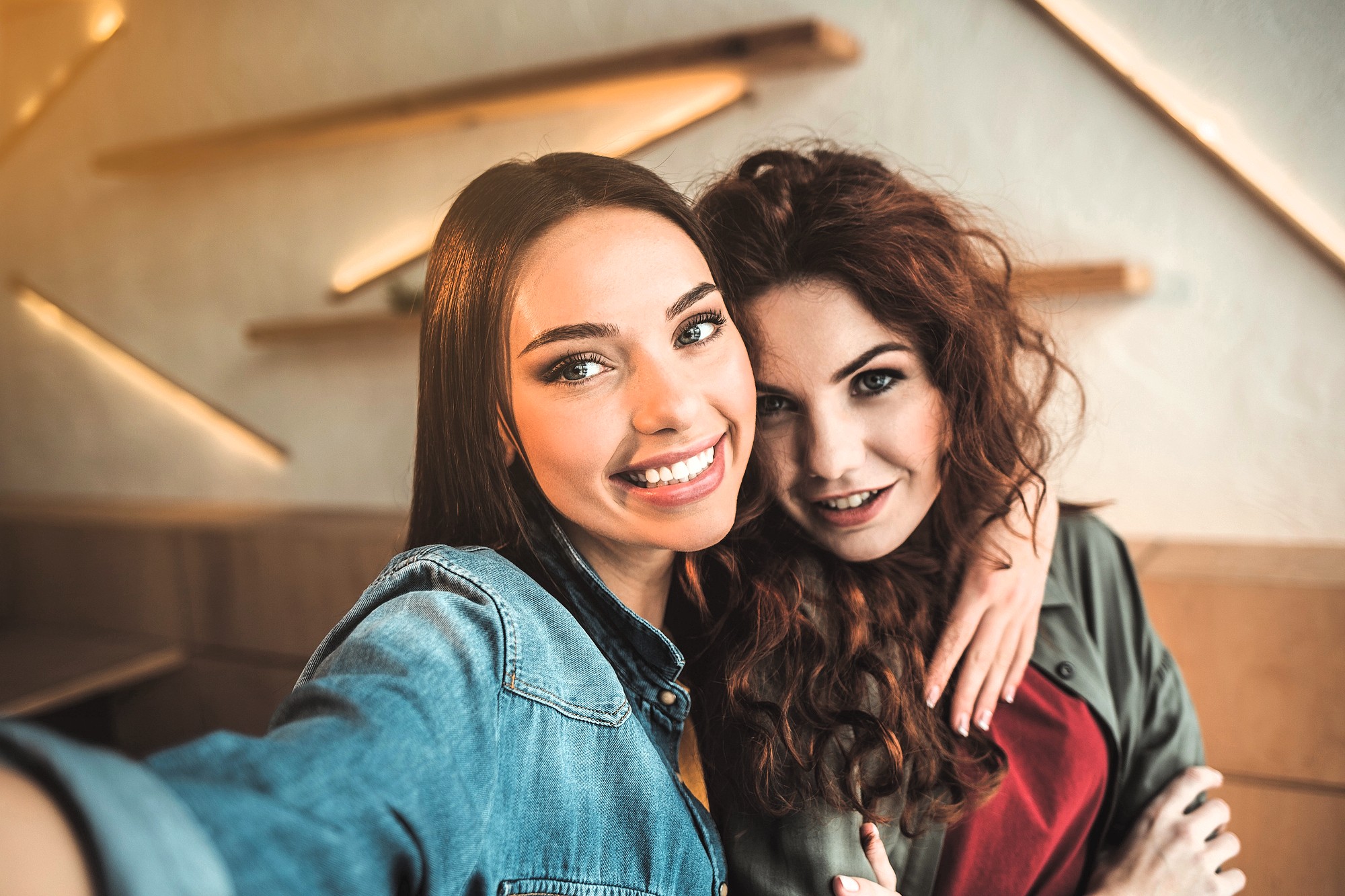 Two people with long hair smile at the camera while taking a selfie indoors. One wears a denim jacket, and the other has a green jacket. The background features angled wooden shelves on a white wall.