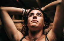 A woman with dark hair reclines, looking upward with arms resting behind her head. She wears a necklace and bracelets, with warm lighting casting soft shadows on her face. The background is blurred and indistinct.