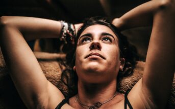 A woman with dark hair reclines, looking upward with arms resting behind her head. She wears a necklace and bracelets, with warm lighting casting soft shadows on her face. The background is blurred and indistinct.