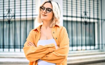 A person with short blond hair and glasses stands confidently, wearing an orange shirt over a white top. They are smiling slightly and have their arms crossed. The background features a window with metal bars.