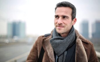 A man with dark hair and a light beard is smiling and looking to the side. He is wearing a brown coat and a gray scarf, standing outdoors with blurred buildings in the background. The scene appears cold and overcast.