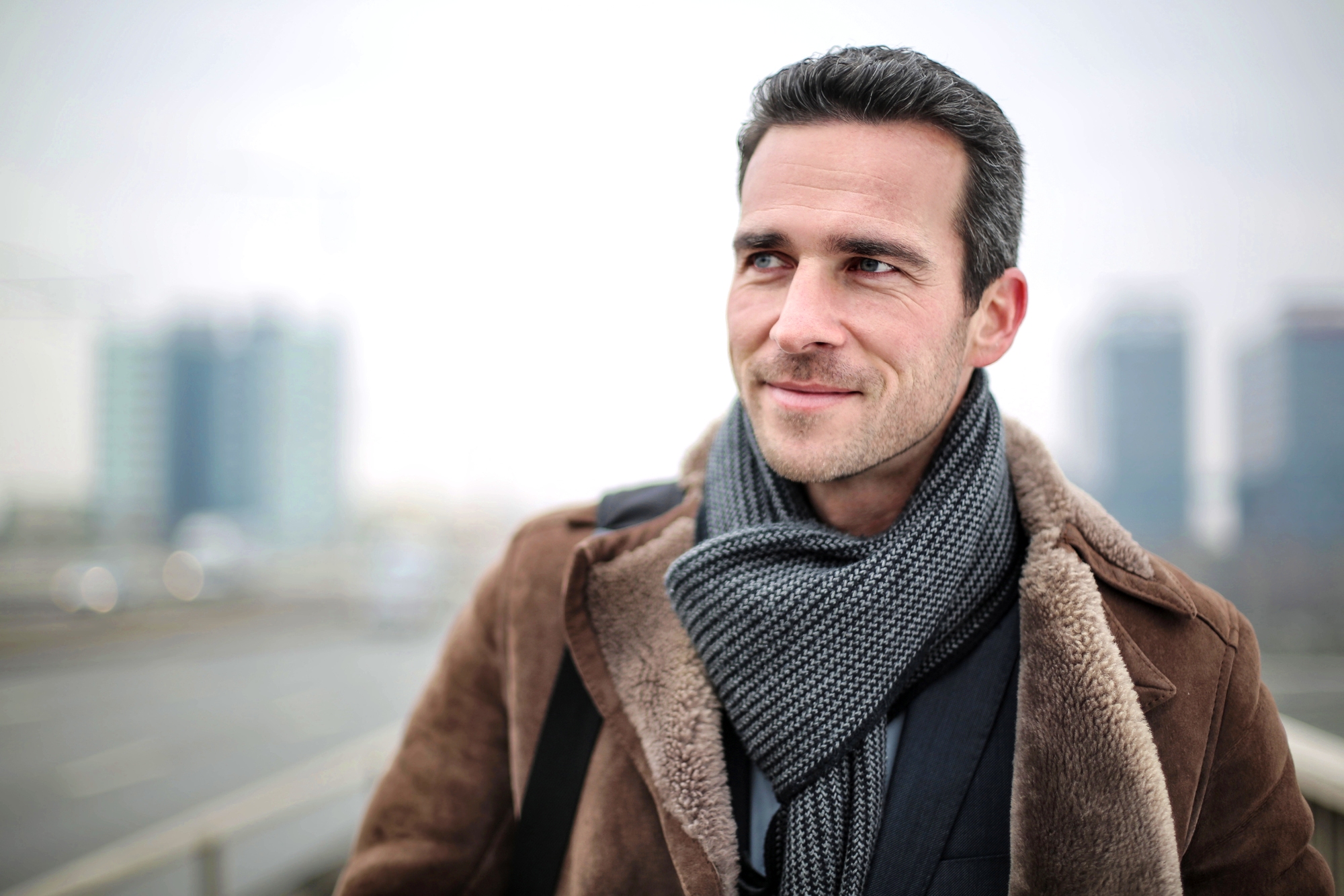 A man with dark hair and a light beard is smiling and looking to the side. He is wearing a brown coat and a gray scarf, standing outdoors with blurred buildings in the background. The scene appears cold and overcast.
