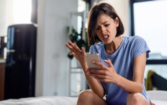 A young woman sitting indoors on a bed looks frustrated while holding a smartphone in one hand and gesturing with the other, her face showing irritation or anger.