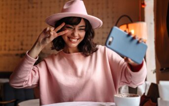 A smiling young woman in a pink sweater and hat takes a selfie with her phone, holding up a peace sign with her fingers, sitting at a table in a cozy café.