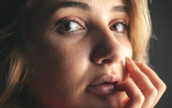 A close-up of a young person with light skin, light brown eyes, and blonde hair. They are looking thoughtfully at the camera, with their fingers gently touching their lips and cheek, in soft natural light.