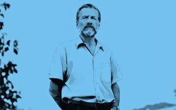 A man with a mustache and beard stands outdoors, wearing a short-sleeve button-up shirt. The image has a blue filter and some trees are visible in the background.