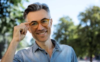 A smiling man with short gray hair and a beard, wearing orange glasses and a light blue denim shirt, stands outdoors in a park. He is touching his glasses with his right hand. The background features blurred green trees and a clear blue sky.