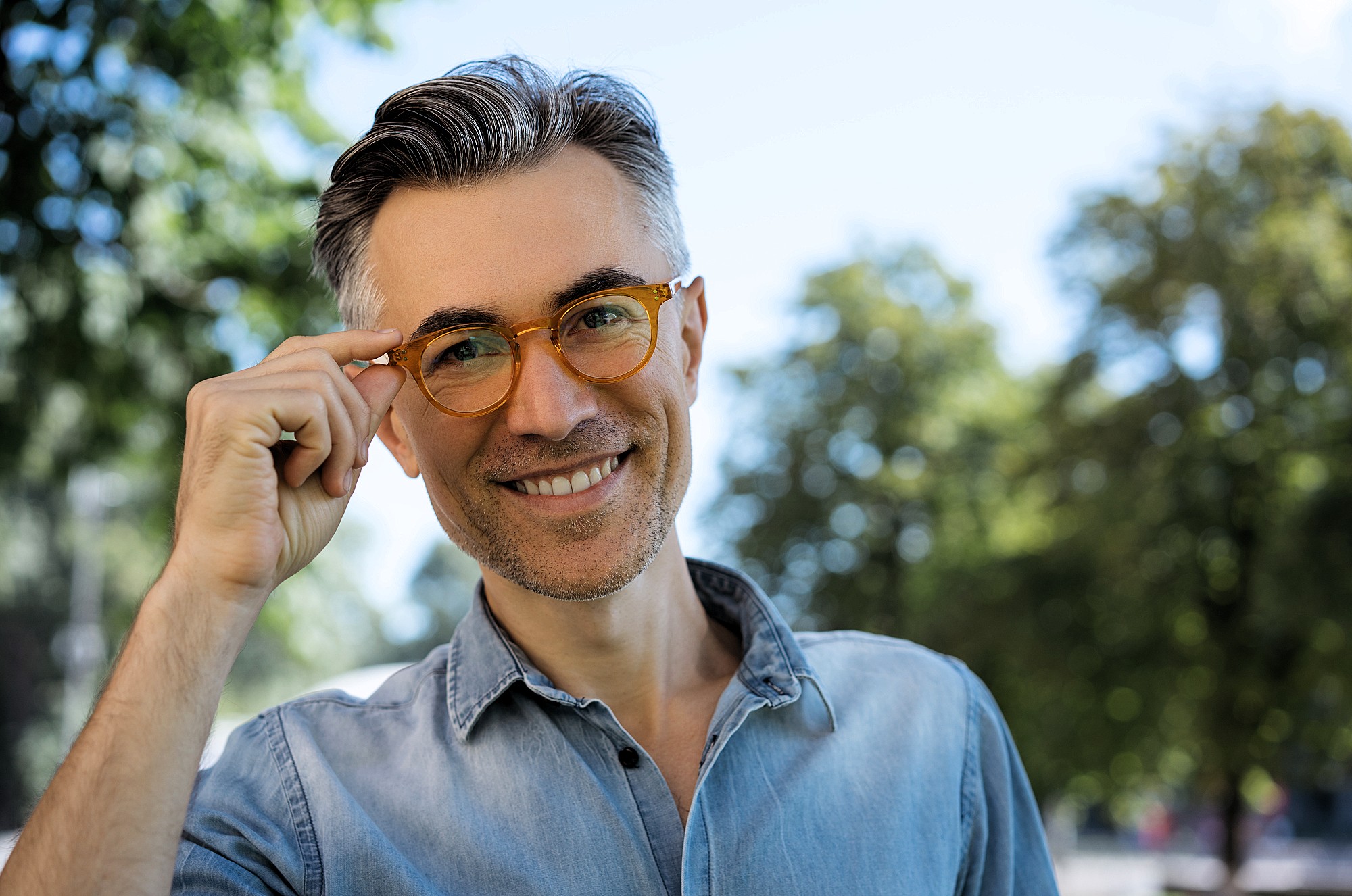 A smiling man with short gray hair and a beard, wearing orange glasses and a light blue denim shirt, stands outdoors in a park. He is touching his glasses with his right hand. The background features blurred green trees and a clear blue sky.
