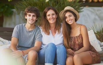A woman sits outdoors on a bench between a young man and a young woman. All three are smiling at the camera. The young woman wears a sunhat, and greenery is visible in the background.