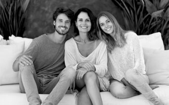 Three people sit together on a couch, smiling warmly at the camera. They appear relaxed and happy, with greenery in the background. The image is in black and white.