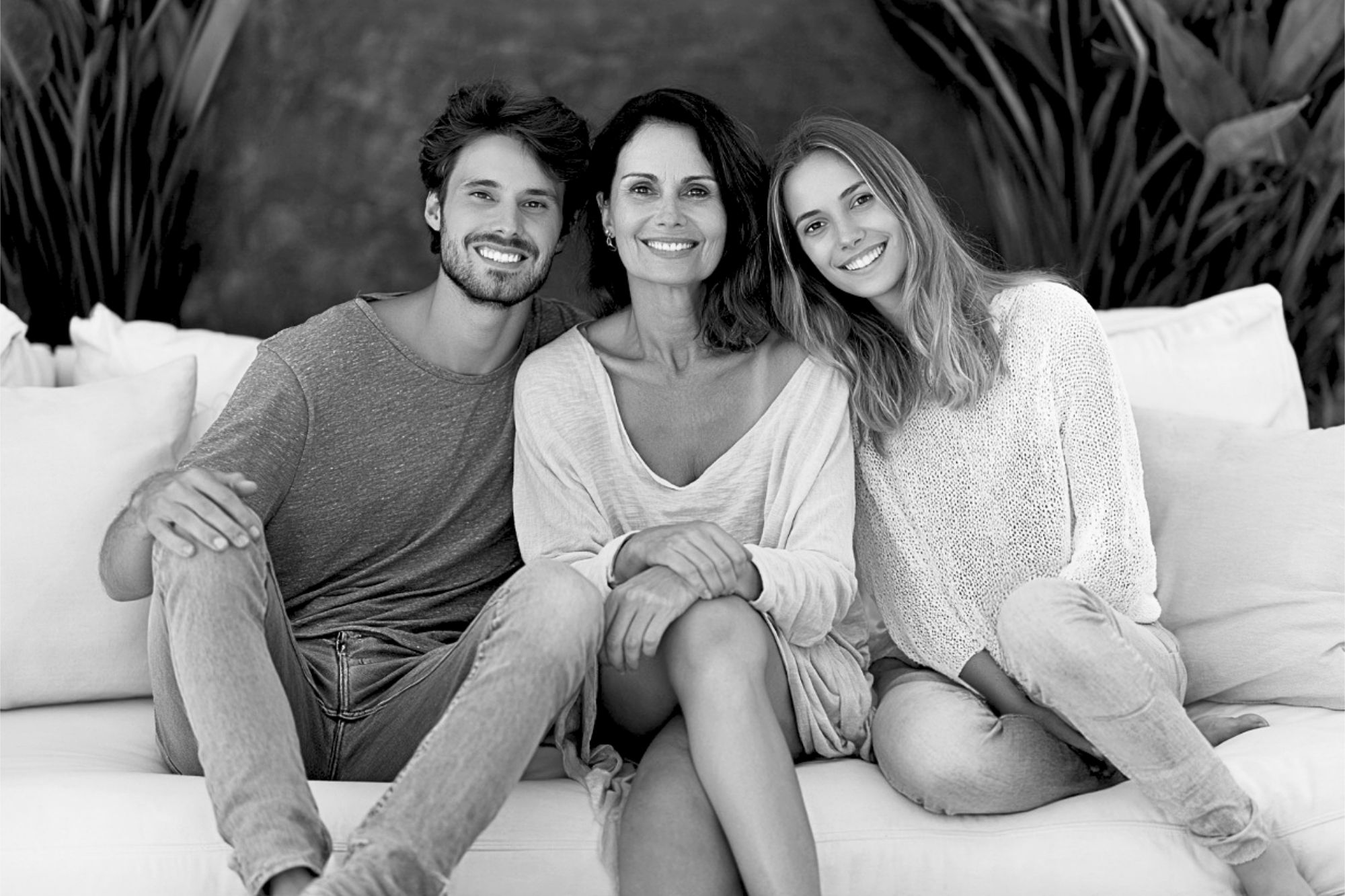 Three people sit together on a couch, smiling warmly at the camera. They appear relaxed and happy, with greenery in the background. The image is in black and white.