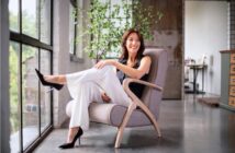 A woman in a black sleeveless top and white pants sits smiling in a modern armchair by large windows, with her legs crossed. There is a plant and wooden furniture visible in the bright, stylish room.