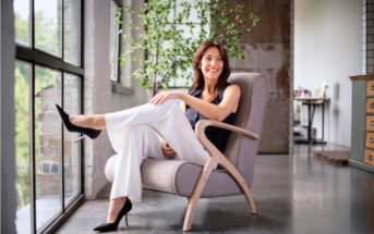 A woman in a black sleeveless top and white pants sits smiling in a modern armchair by large windows, with her legs crossed. There is a plant and wooden furniture visible in the bright, stylish room.