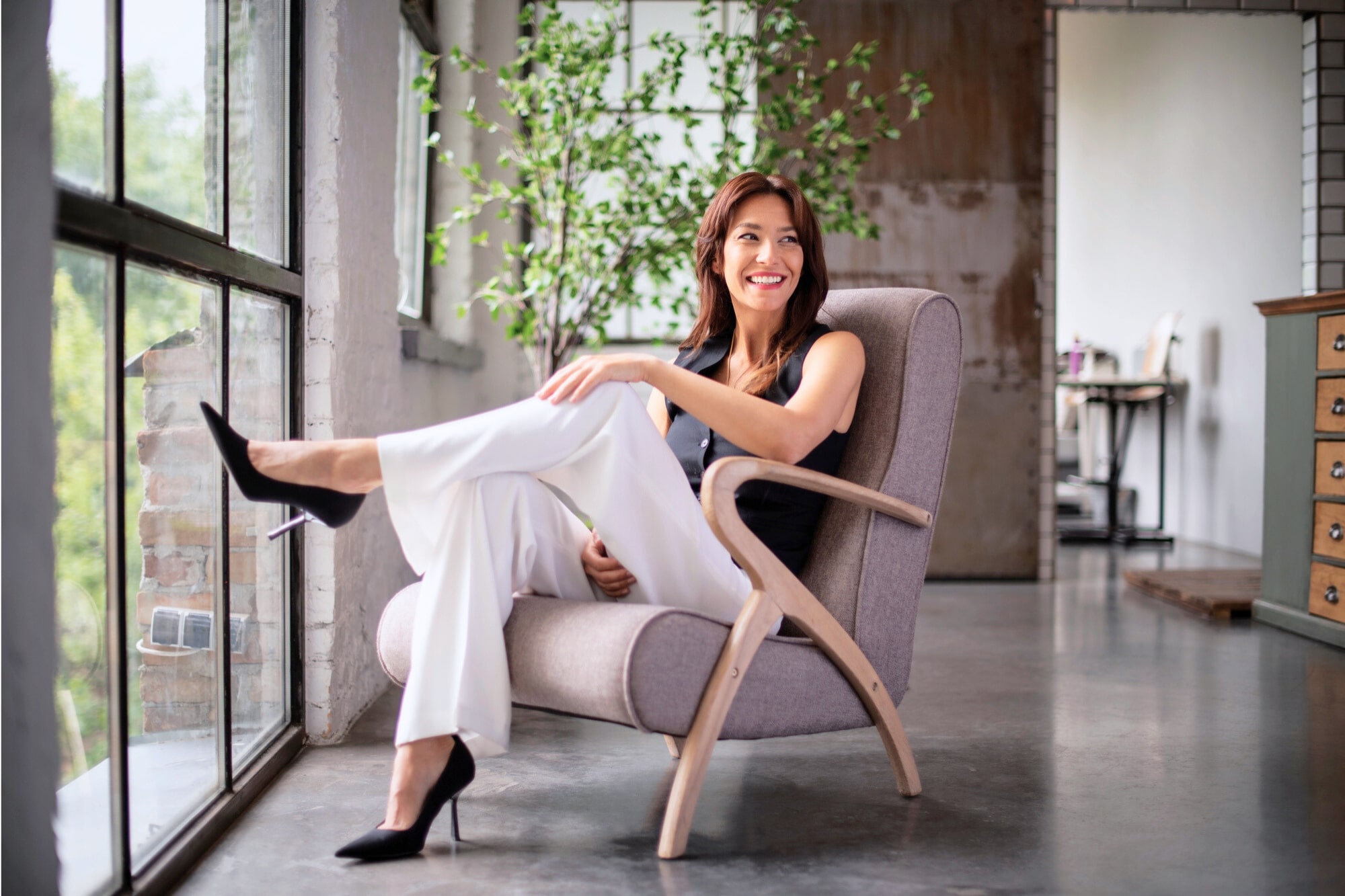 A woman in a black sleeveless top and white pants sits smiling in a modern armchair by large windows, with her legs crossed. There is a plant and wooden furniture visible in the bright, stylish room.