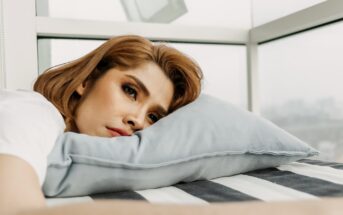 A woman with short brown hair lies on a striped bed, resting her head on a light blue pillow. She gazes into the distance with a thoughtful expression. The background shows a window with a blurred view outside.