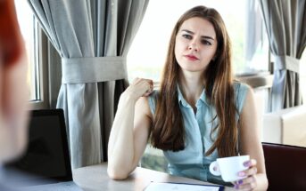 A woman with long brown hair sits at a table with a cup in one hand, looking thoughtfully at someone off-camera. A tablet with a colorful chart and a laptop are on the table in front of her.
