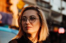 A person with short blonde hair, wearing round glasses and a nose ring, looks thoughtfully into the distance. The background shows an urban street scene bathed in warm sunlight.