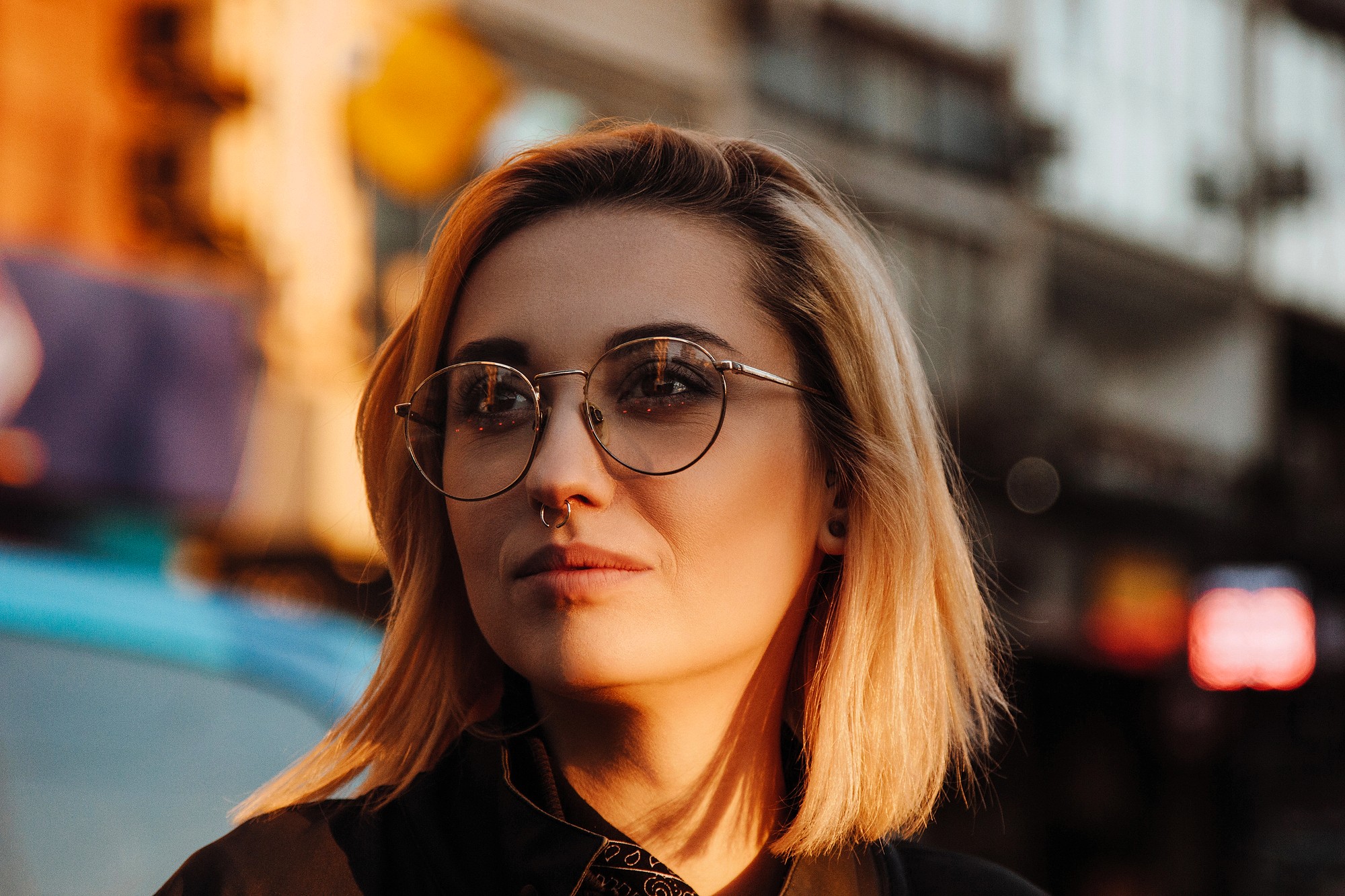 A person with short blonde hair, wearing round glasses and a nose ring, looks thoughtfully into the distance. The background shows an urban street scene bathed in warm sunlight.